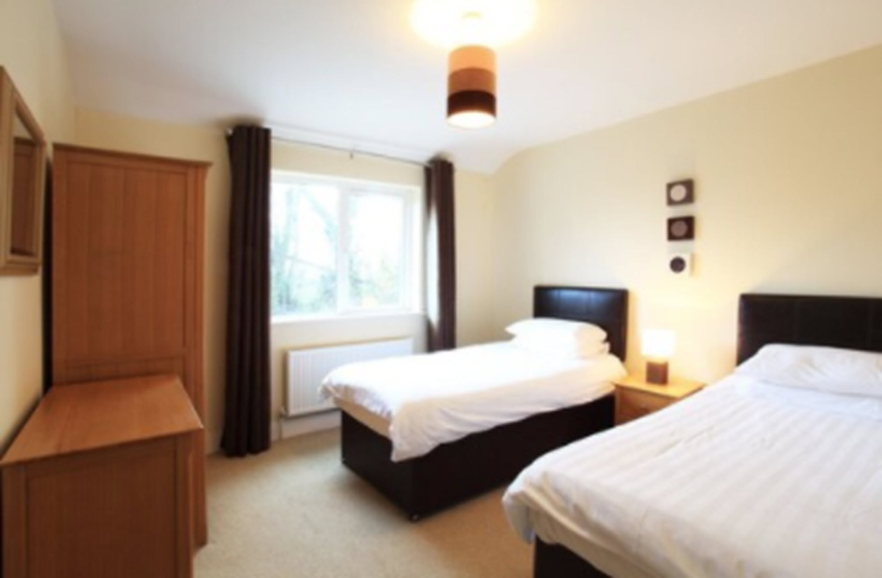 A hotel room with two beds, a wooden wardrobe, a window with curtains, and a wall-mounted light fixture.