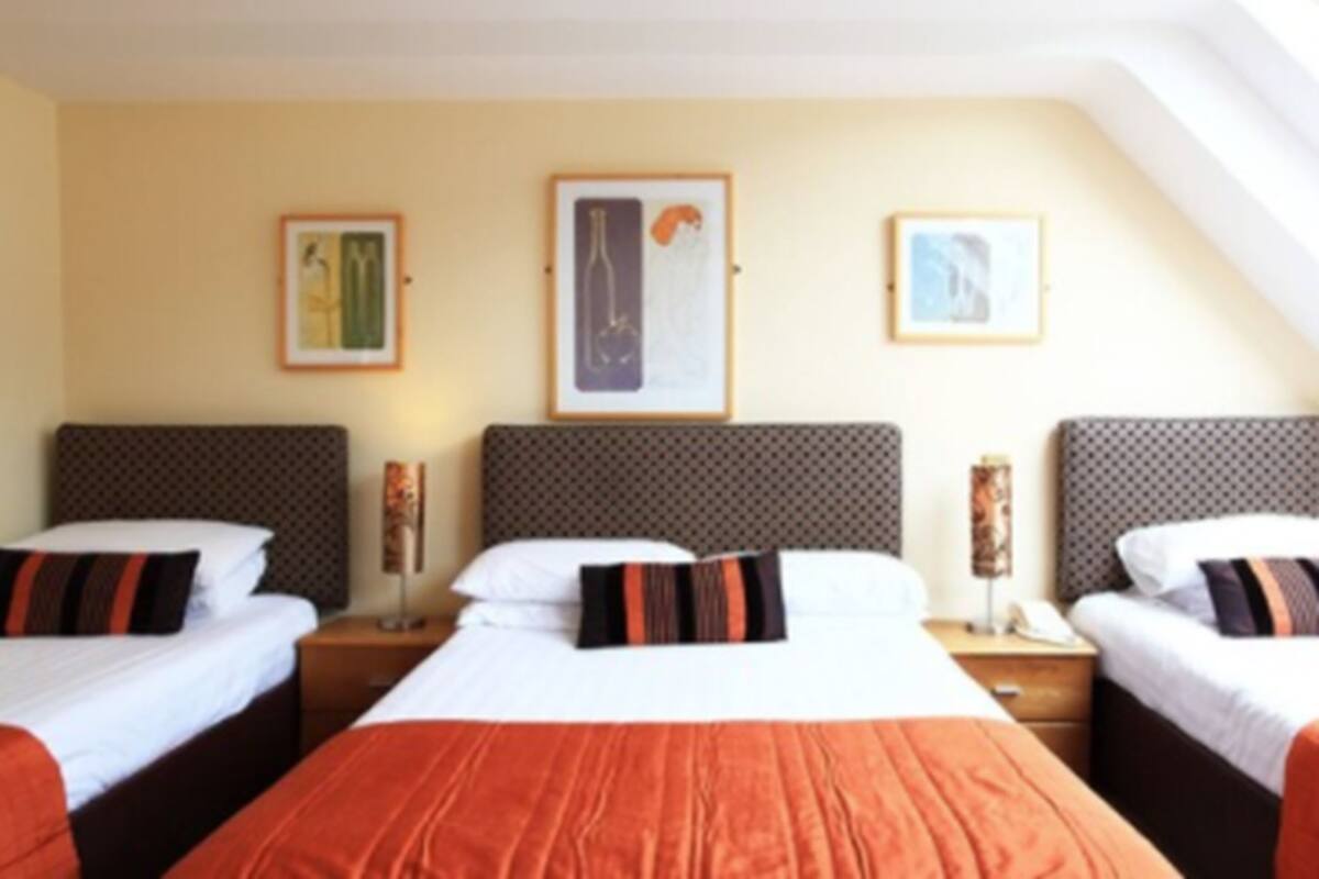 Een hotelkamer met drie bedden, elk voorzien van een gestreept hoofdeinde en kussens. Aan de muur boven de bedden hangen kunstwerken in lijsten.