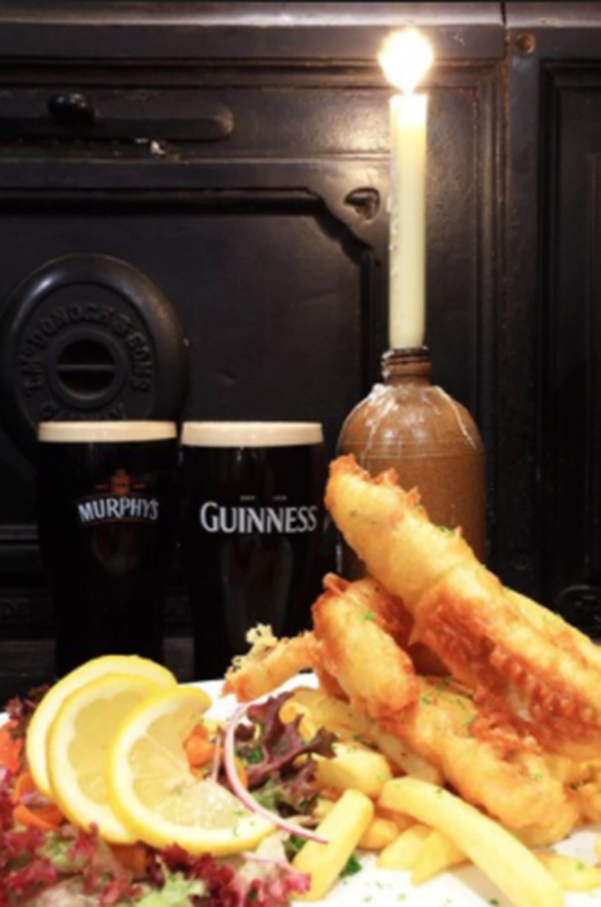 Un plato de pescado y papas fritas con rodajas de limón y una ensalada, acompañado por dos jarras de cerveza con las etiquetas 'Murphy's' y 'Guinness'.