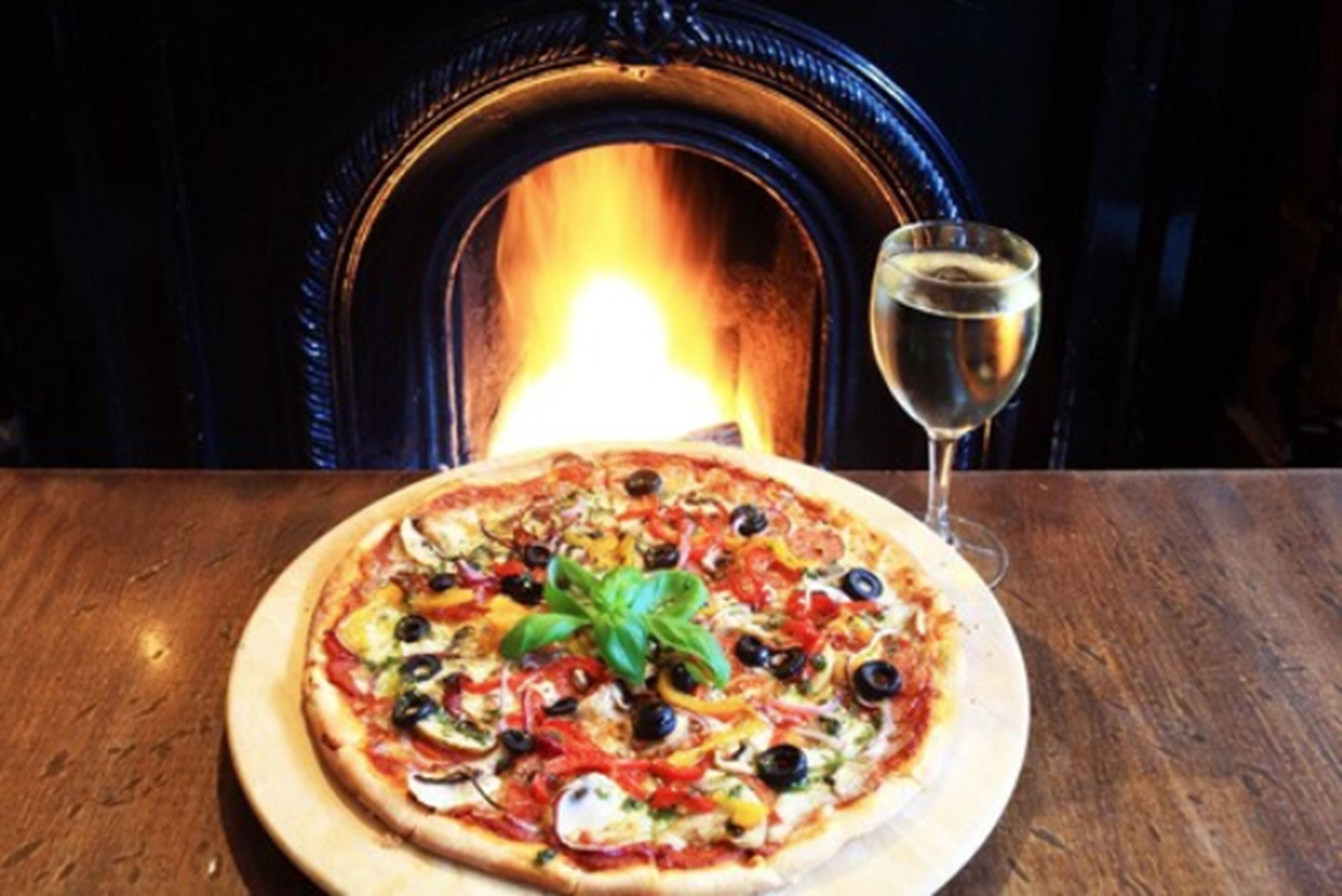 A pizza on a wooden table with a glass of white wine and a fireplace in the background.