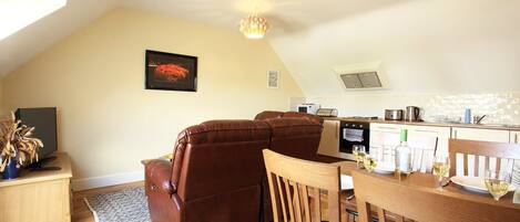 A cozy living room with a brown leather sofa, a wooden dining table with chairs, a kitchen area with a microwave and stove, and a framed picture on the wall.