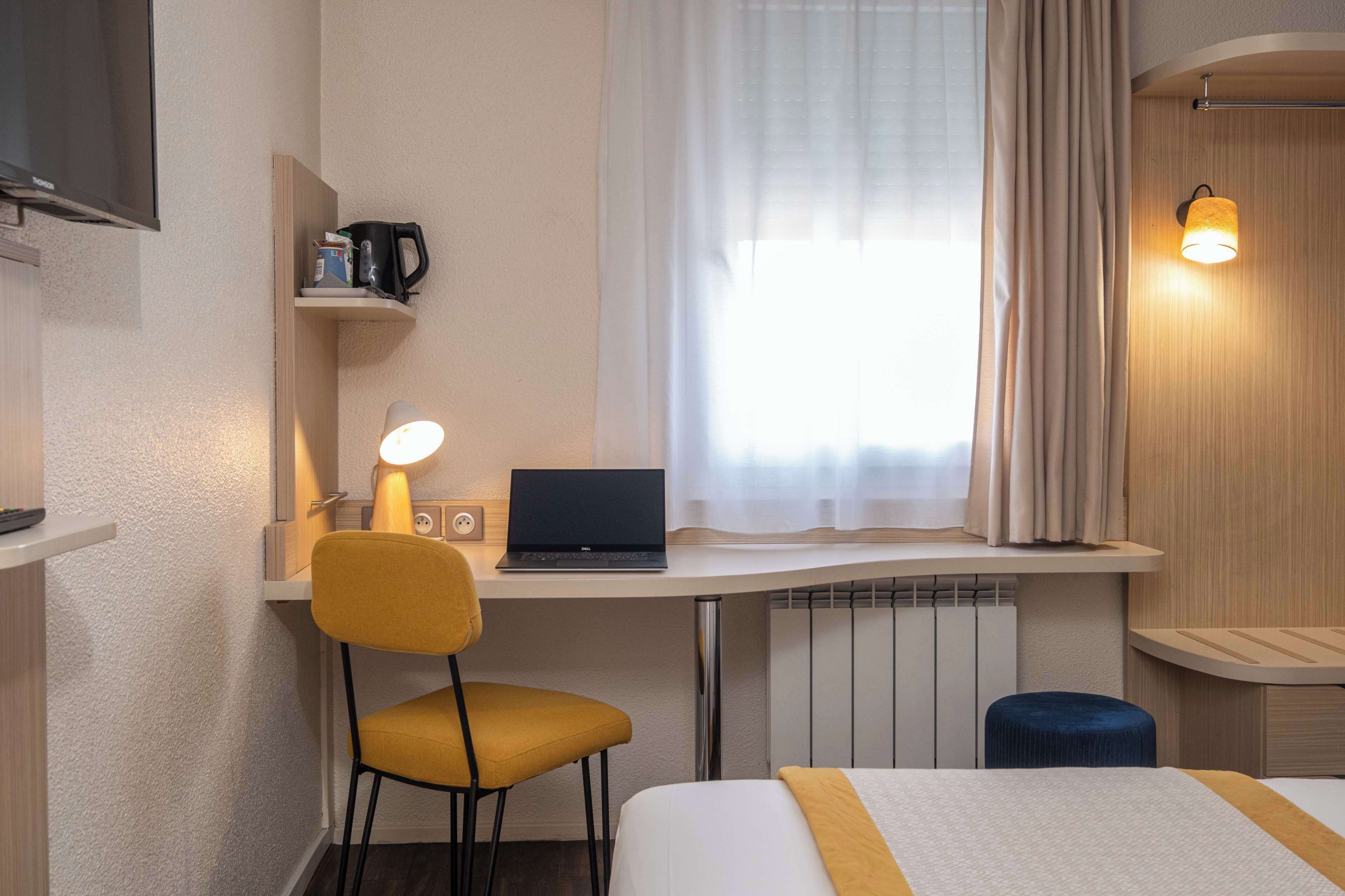 Habitación de hotel con cama, escritorio con laptop, silla, taburete pequeño, estante con cafetera y televisor montado en la pared.