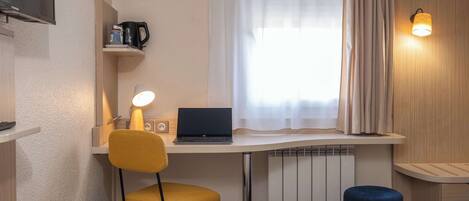 Habitación de hotel con cama, escritorio con laptop, silla, taburete pequeño, estante con cafetera y televisor montado en la pared.