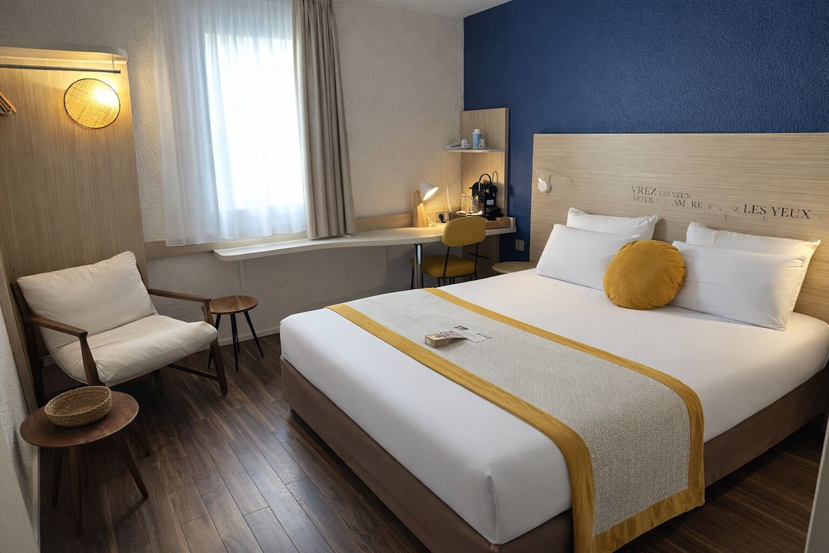 Une chambre d’hôtel avec un lit, une chaise, un bureau et une fenêtre avec des rideaux.