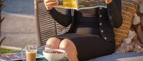 A person sitting on a wicker chair reading a magazine, with a glass of coffee and a bowl of cereal on a small table beside them.