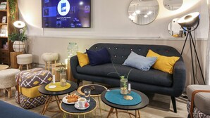 Ein modernes Wohnzimmer mit einem dunkelgrauen Sofa, gelb-blauen Kissen, runden Tischen mit Getränken und Snacks sowie einem Fernseher, der ein Programm zeigt.