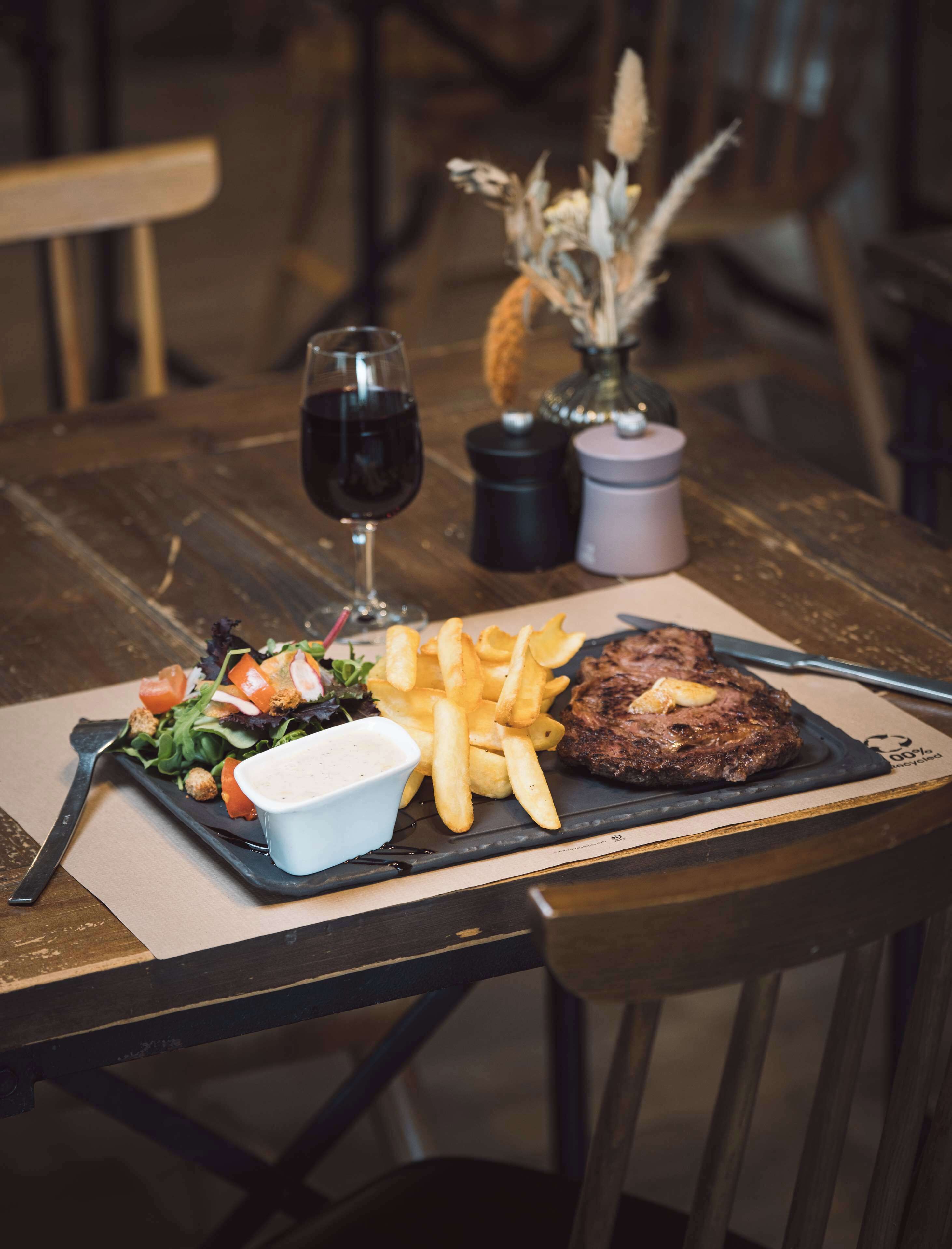 A meal consisting of a steak, fries, salad, and a glass of red wine on a wooden table.