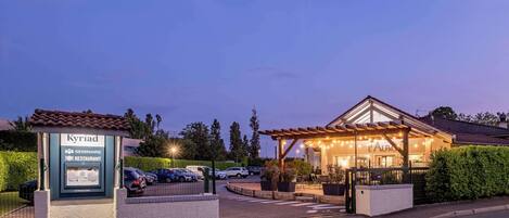 A hotel entrance with a signboard, a covered outdoor seating area, and a parking lot.