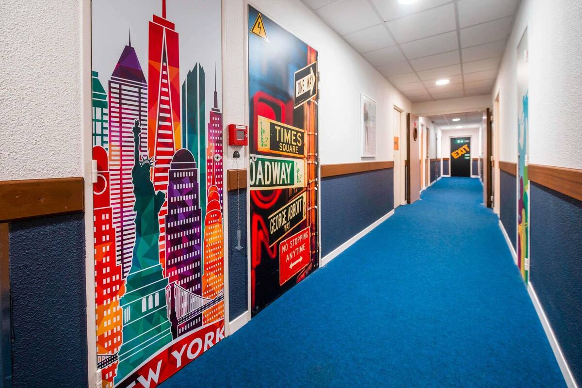 A hotel corridor with a blue carpet, walls decorated with colorful cityscape murals, and directional signs for Times Square and Broadway.