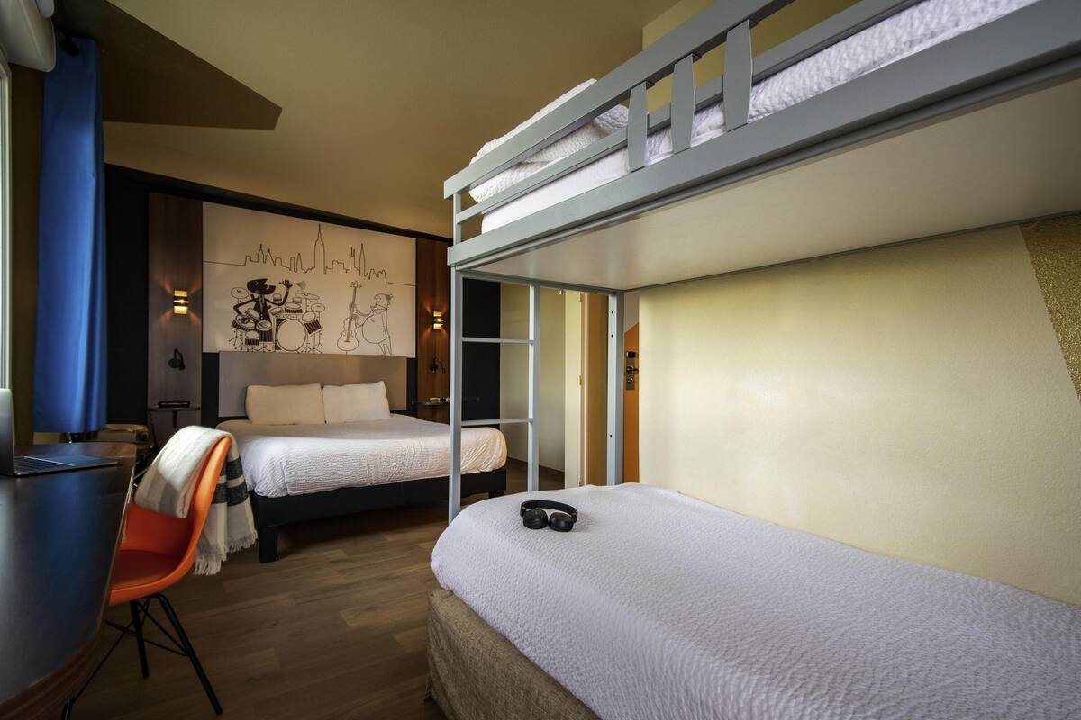 A hotel room with a bunk bed, a desk with a laptop, a chair, and a bed with a blanket.