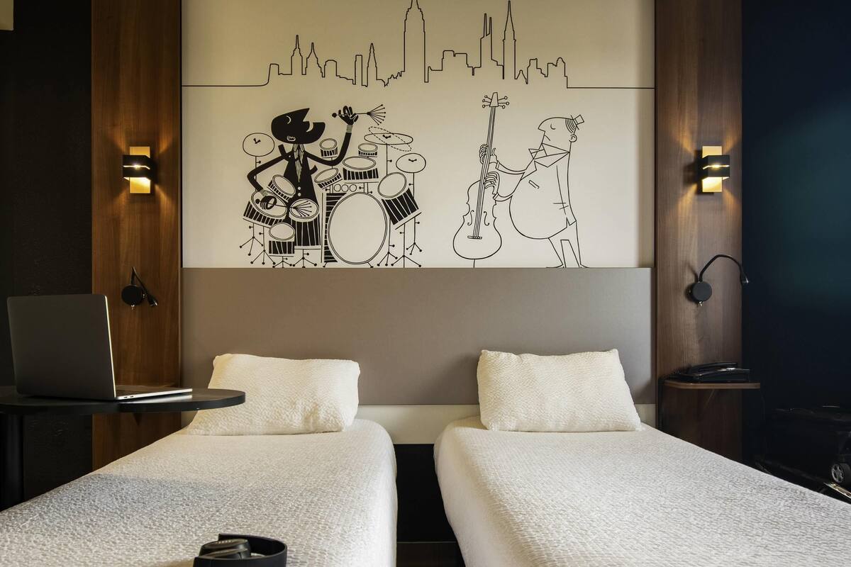A hotel room with two single beds, a laptop on a side table, and a wall mural of musicians.