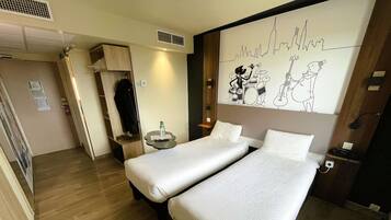 Habitación de hotel con dos camas, una mesita con una botella, perchero y un cuadro de un horizonte urbano.