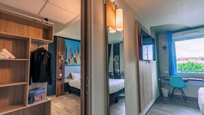 A hotel room with a wooden wardrobe, a bed with a cityscape mural, a desk with a chair, and a window with a view of buildings. - ibis Styles Toulouse Blagnac Aeroport (Blagnac)