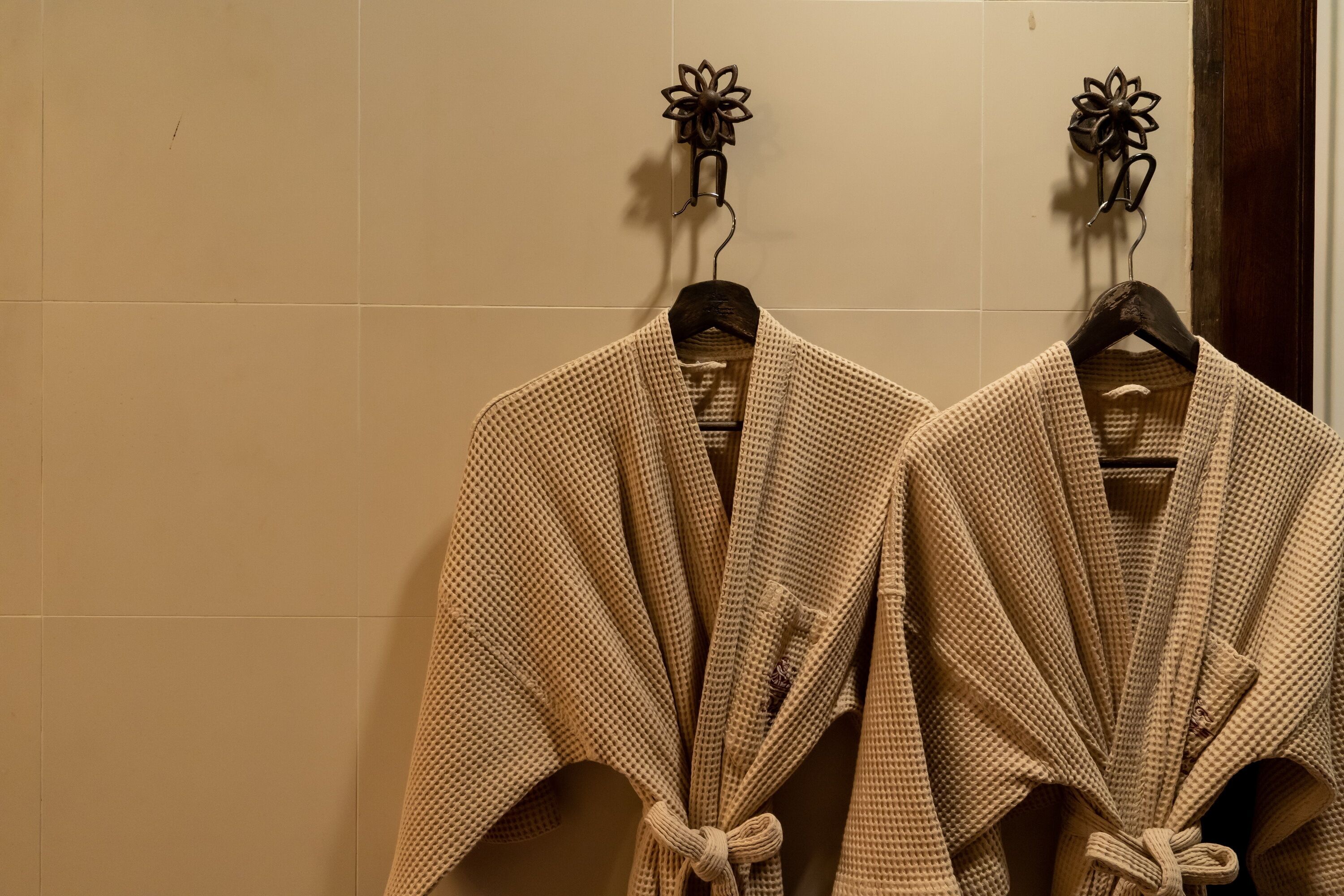 Two beige robes tied on wooden hangers against a tiled wall.