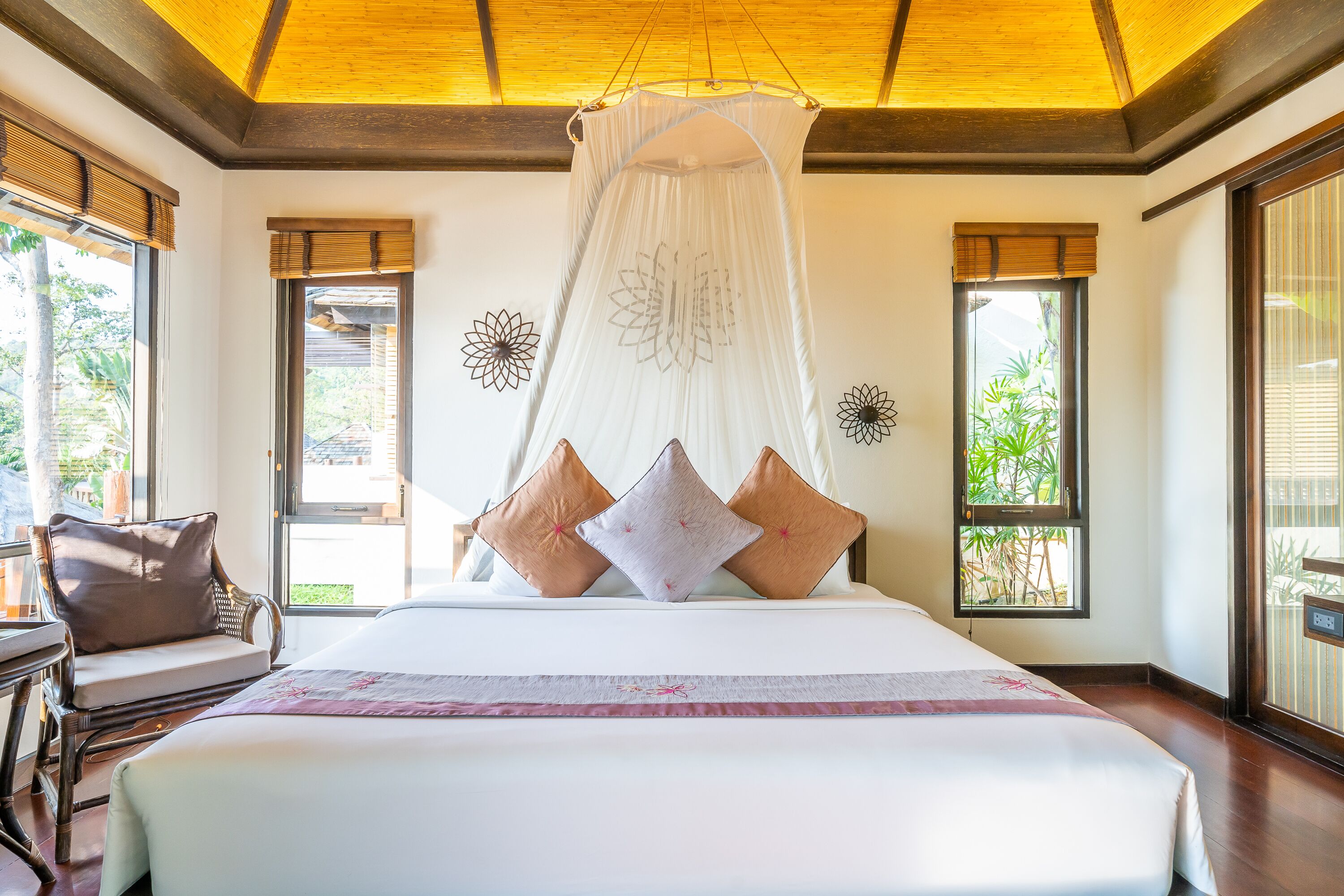 A large bed with a canopy, a chair, and a view of greenery outside.
