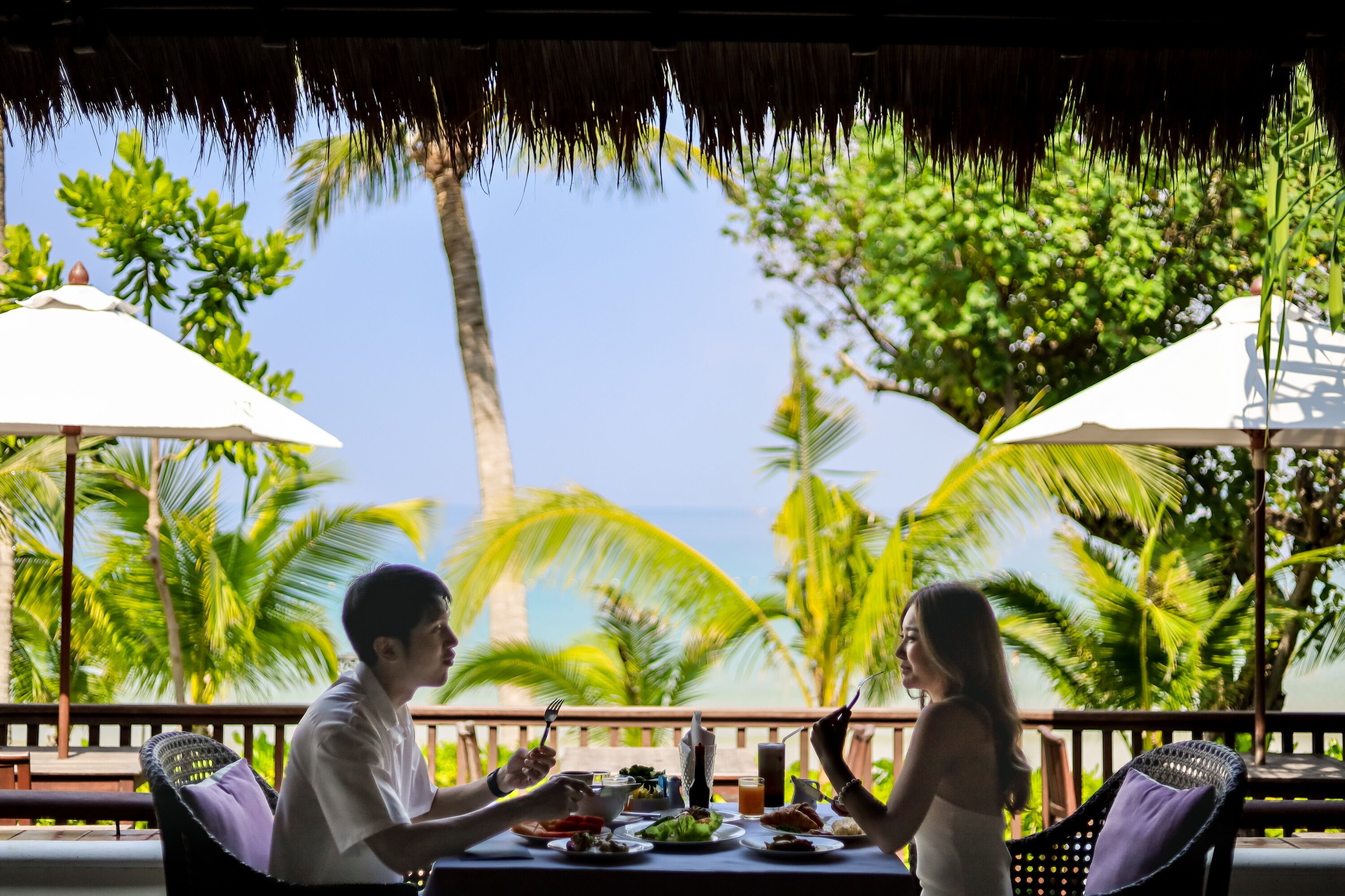 A dining table with food and drinks, surrounded by chairs, under a thatched roof with palm trees in the background.