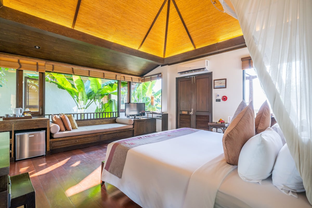 A bedroom with a bed, bedside table, and a window with a view of greenery.