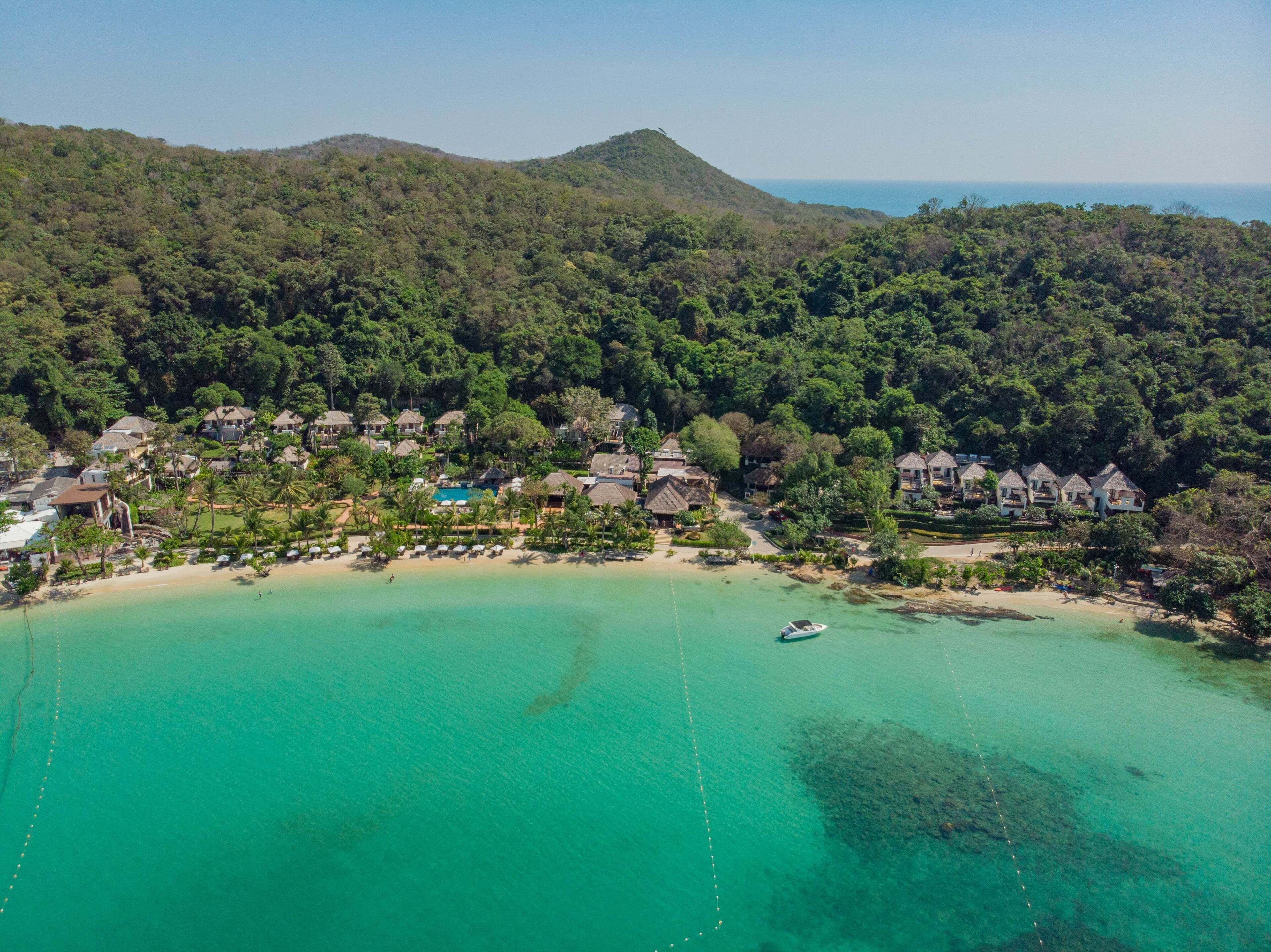 An aerial view of a coastal resort with beachfront villas, a swimming pool, and a boat on the water.