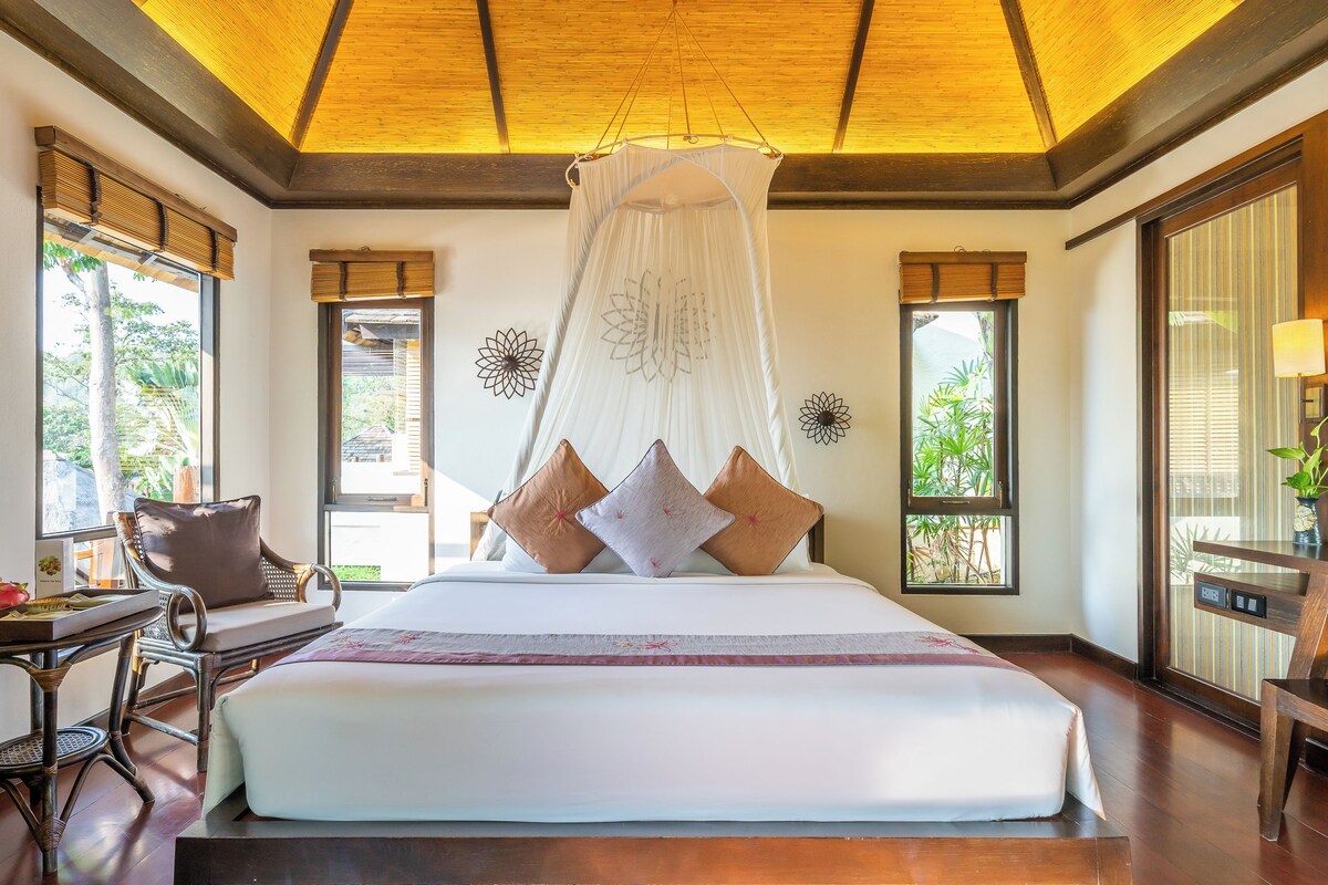 Une chambre spacieuse avec un grand lit, un baldaquin et une vue sur la verdure par les fenêtres.