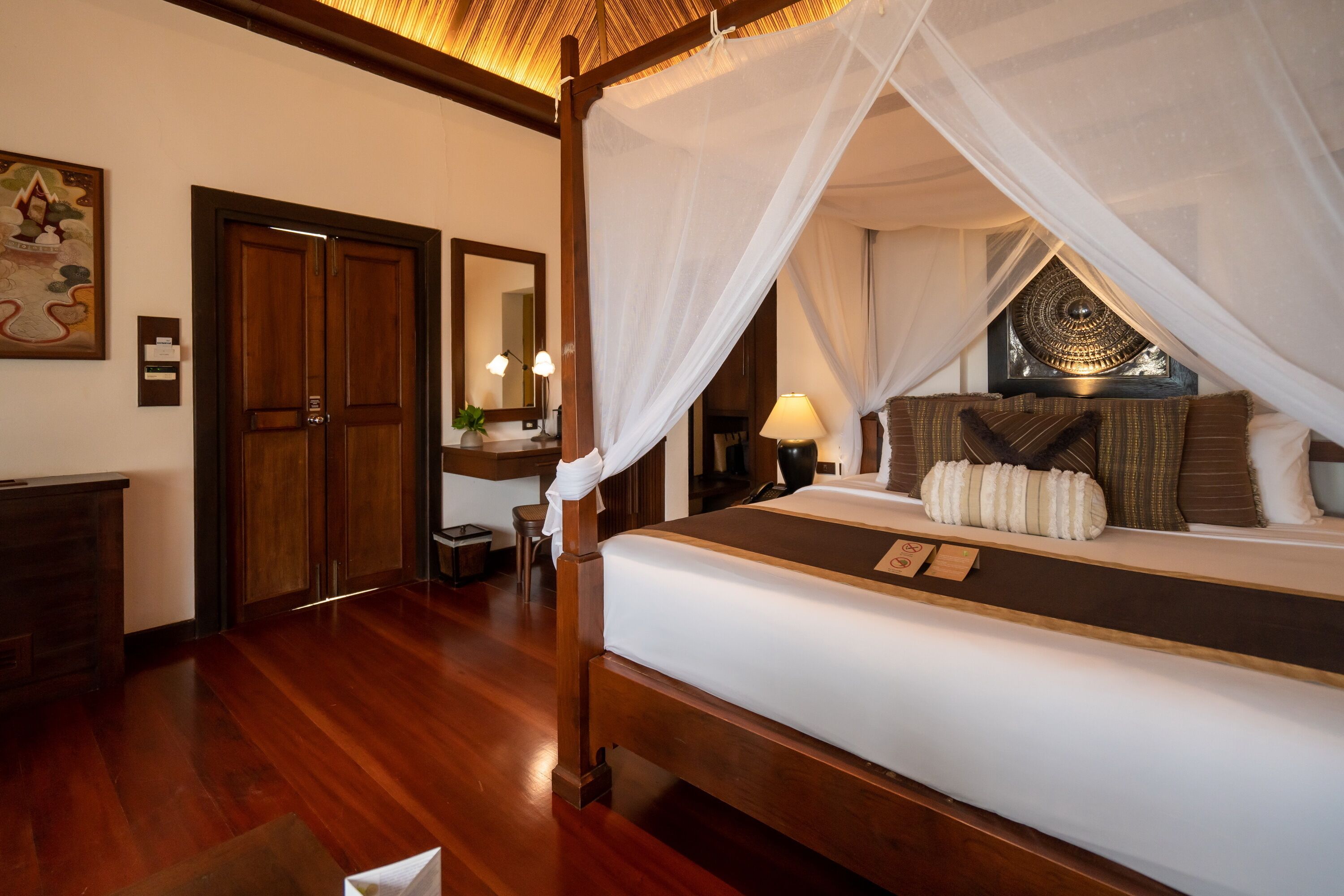 A four-poster bed with a canopy, wooden flooring, a desk with a chair, a mirror, and a framed picture on the wall.