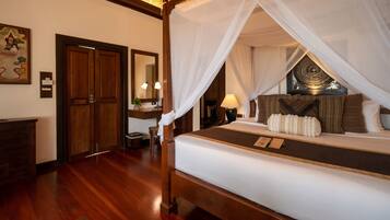 A four-poster bed with a canopy, wooden flooring, a desk with a chair, a mirror, and a framed picture on the wall.