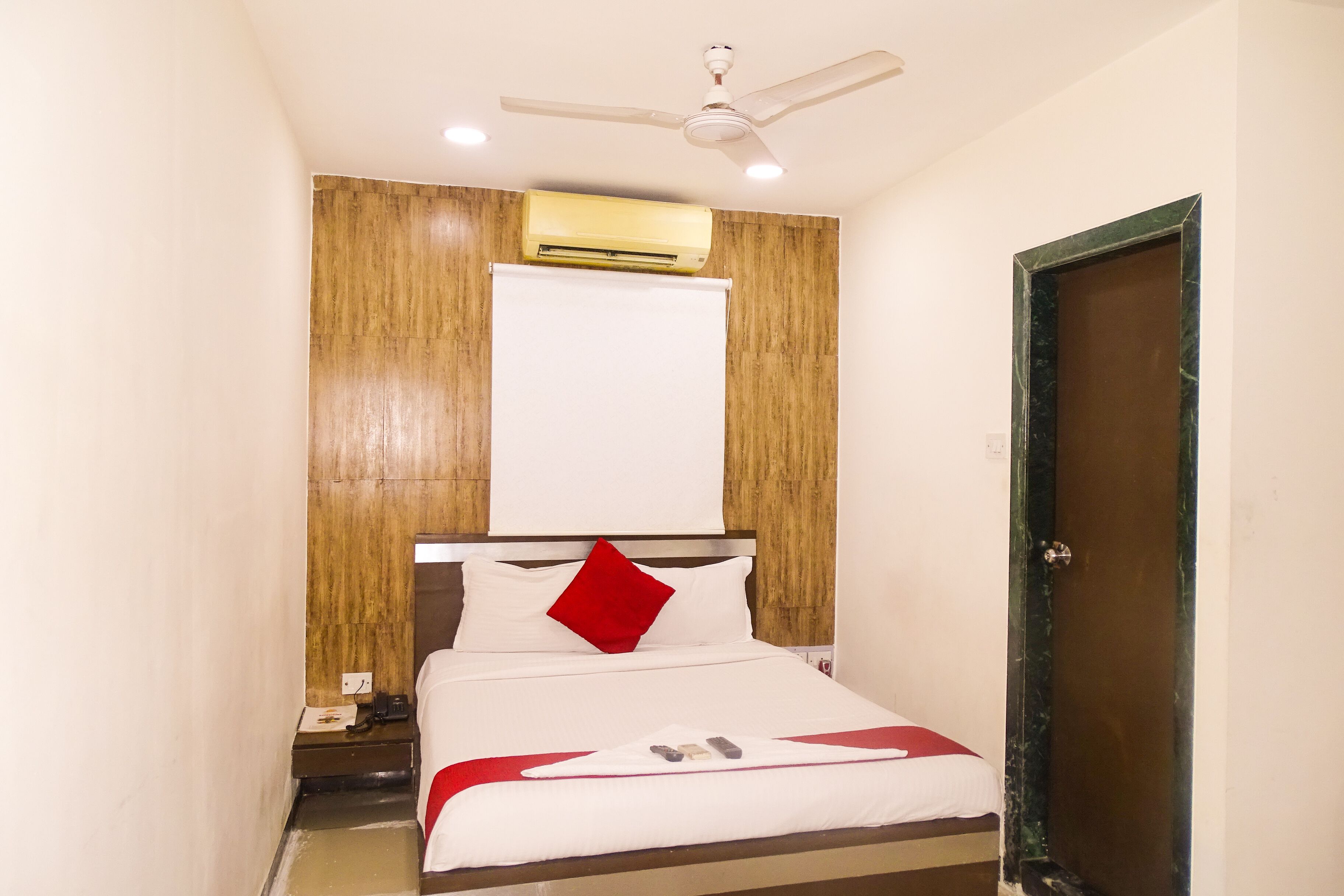 Hotelkamer met een bed, plafondventilator, airconditioning, houten paneelwand, een witte deur en nachtkastje met telefoon.