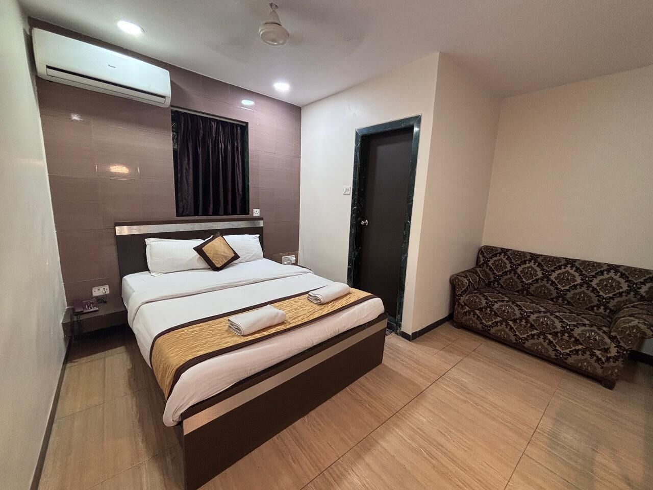A hotel room with a bed, a sofa, an air conditioning unit, and a door.