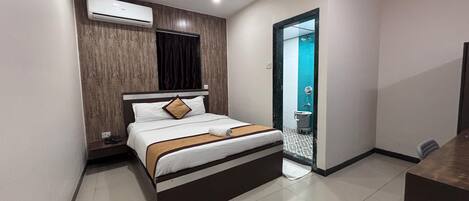 A modern hotel room with a bed, an air conditioning unit, a bathroom with a toilet and sink, and a small desk.