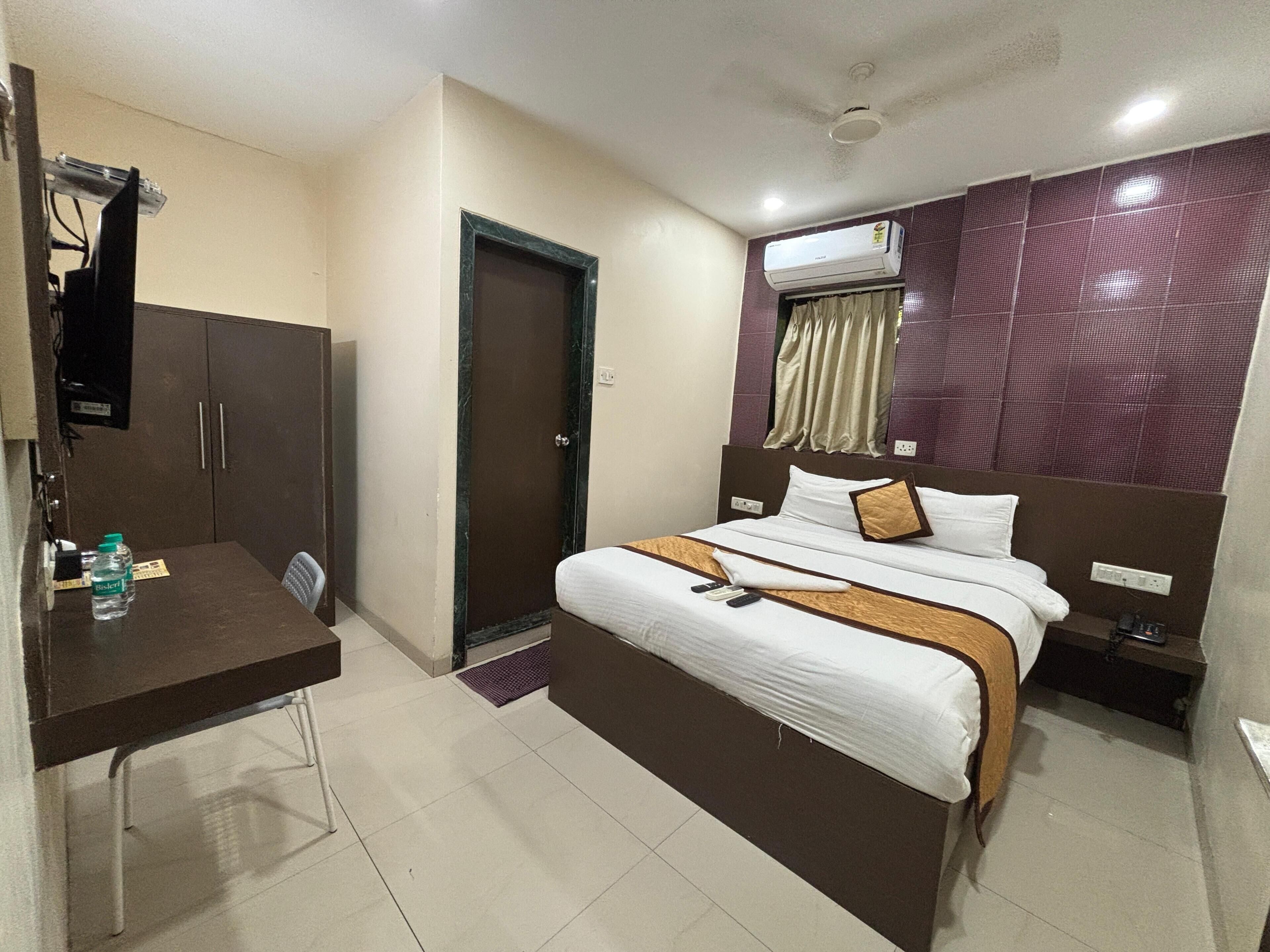 A hotel room with a bed, a desk, a wardrobe, an air conditioning unit, and a door.