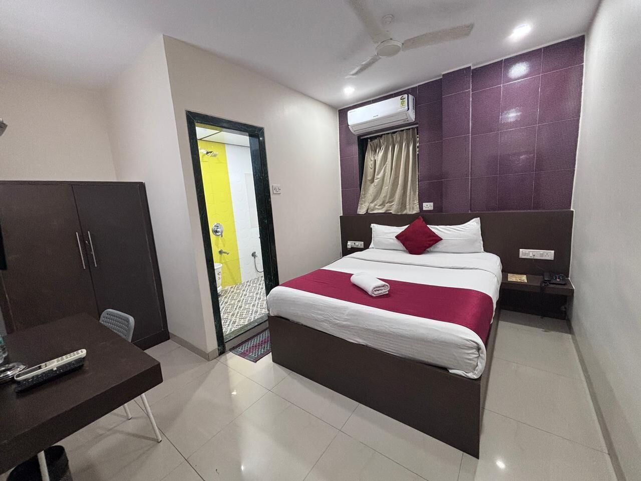 A hotel room with a bed, a desk, a wardrobe, an air conditioning unit, and a bathroom door.