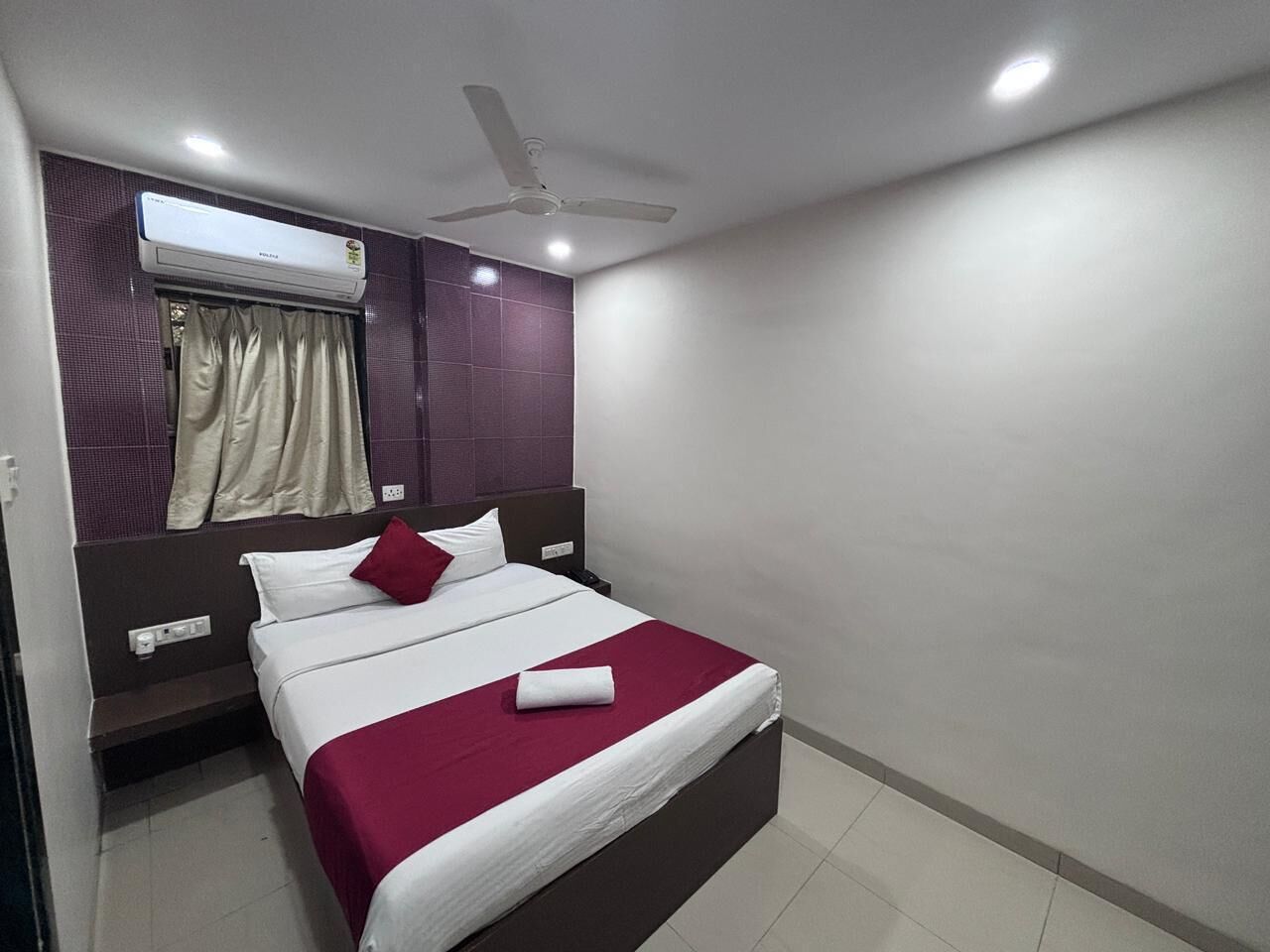 A hotel room with a bed, a ceiling fan, an air conditioning unit, a window with curtains, and a wall-mounted nightstand.