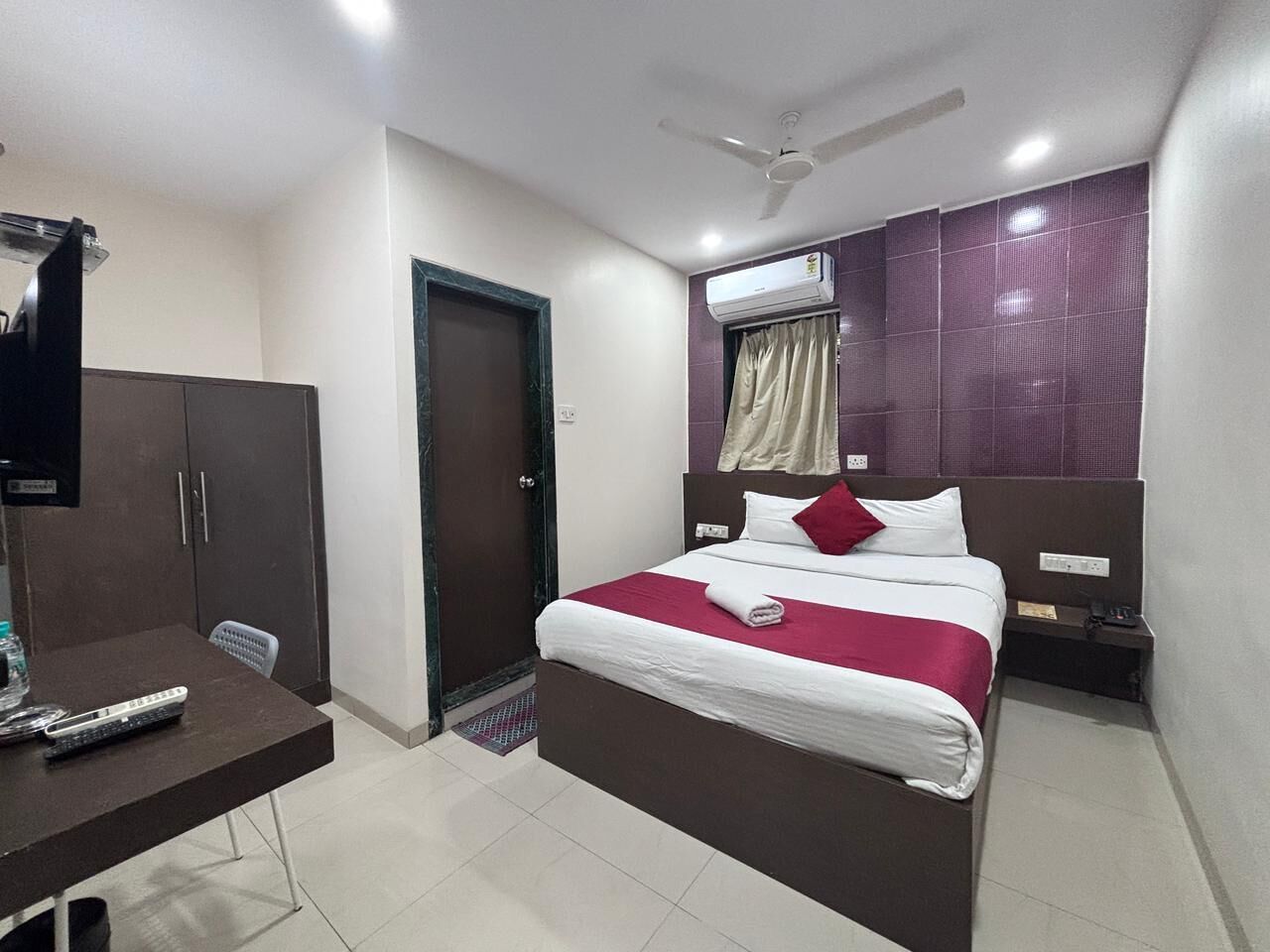 A hotel room with a bed, a desk, a wardrobe, a door, an air conditioning unit, and a ceiling fan.