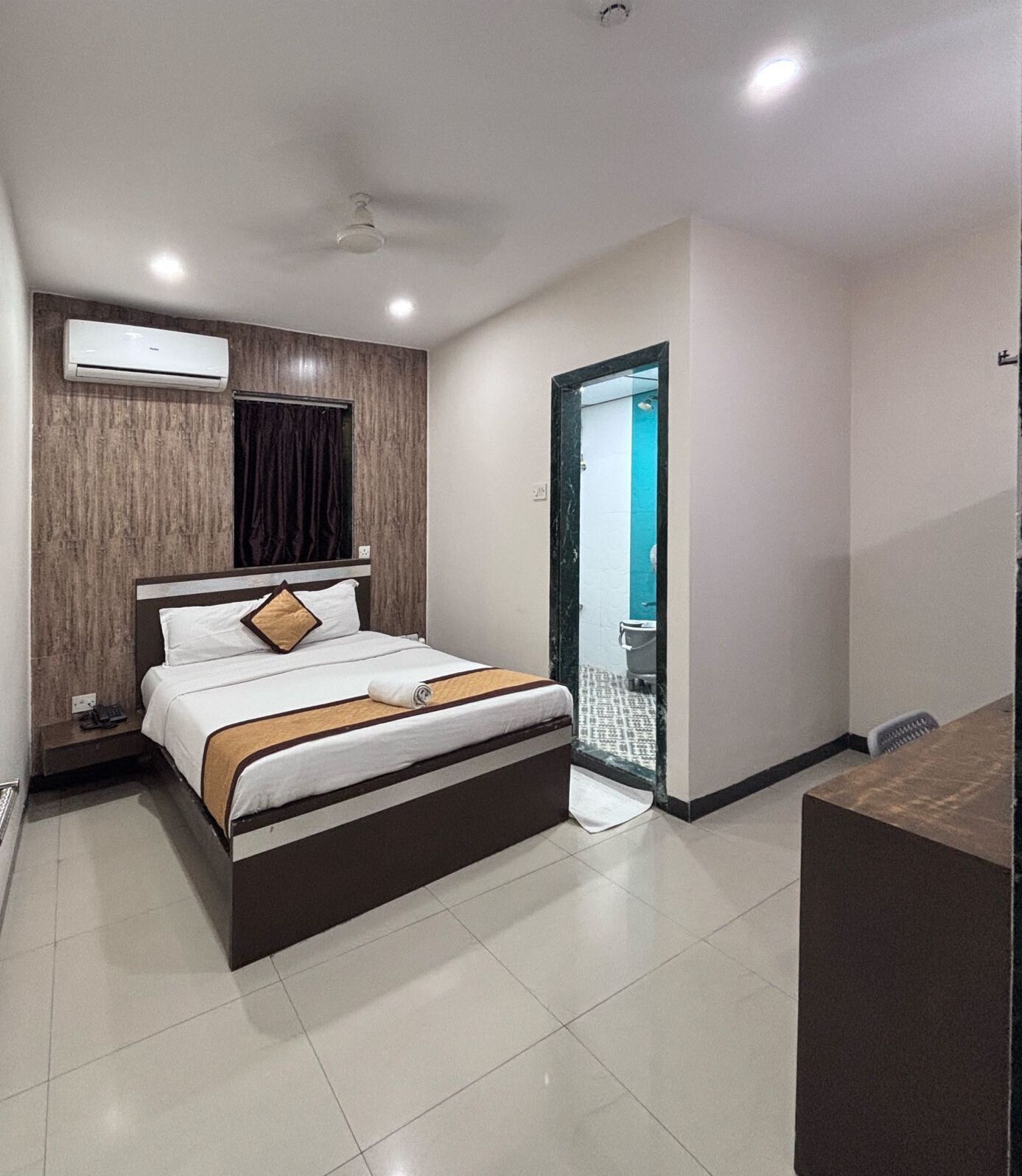A modern hotel room with a bed, bedside table, air conditioning unit, and a bathroom visible through an open door.