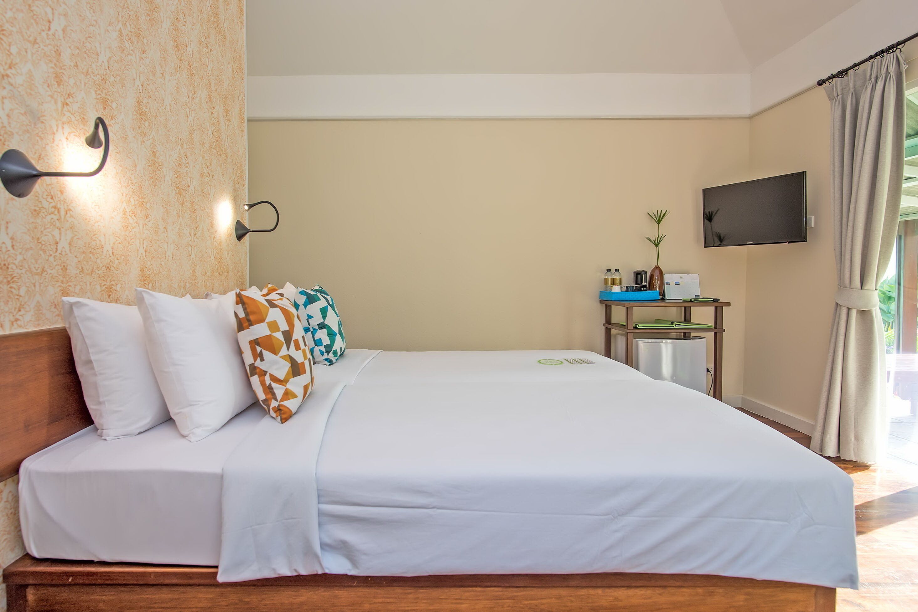 A neatly made bed with white bedding and patterned pillows, a bedside table with a small plant, a flat-screen TV mounted on the wall, and a window with curtains.