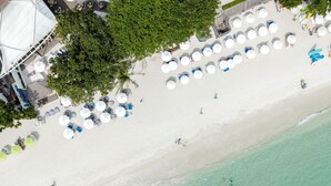 An aerial view of a beach with rows of umbrellas and lounge chairs, a sandy shoreline, and a few people in the water.