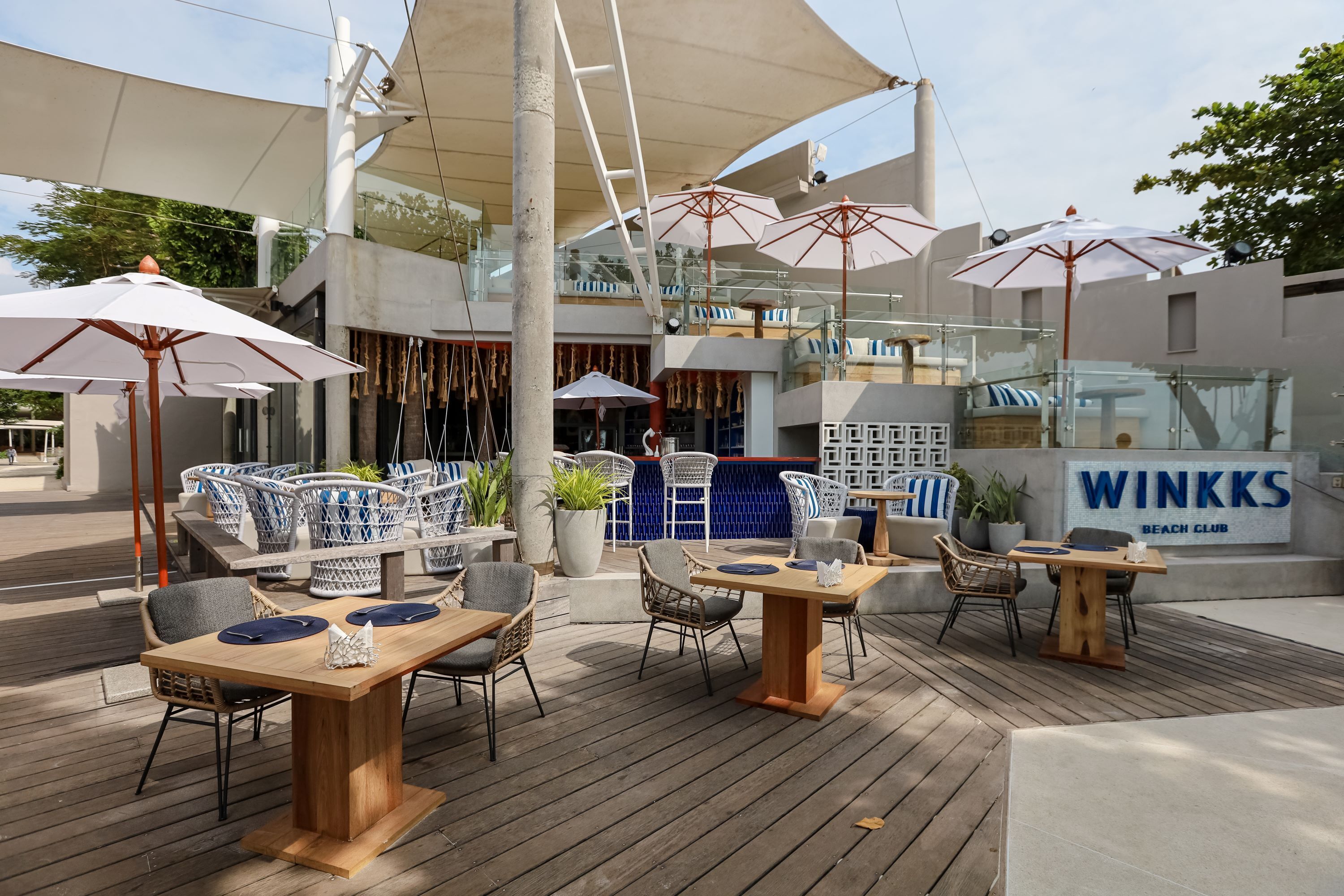 An outdoor seating area with wooden tables and chairs, umbrellas, and a sign reading "WINKKS BEACH CLUB".