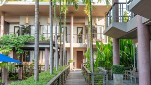 A wooden walkway leading to a building with multiple balconies, surrounded by lush greenery and palm trees.