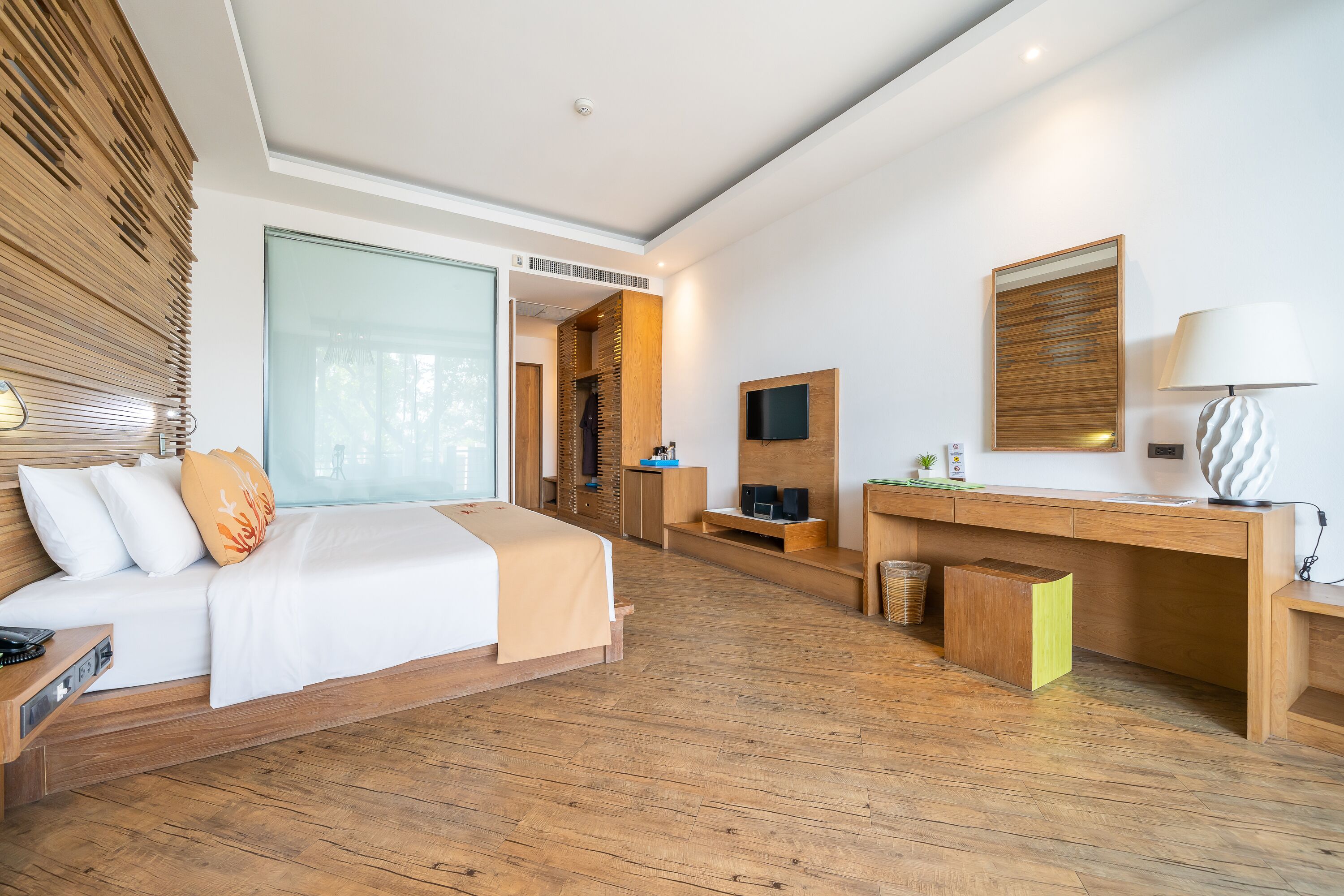 Quarto de hotel moderno com piso de madeira, cama com roupa de cama branca, escrivaninha com luminária e televisão na parede.