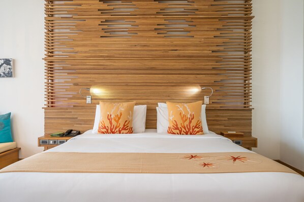 A hotel room with a wooden headboard, a bed with white bedding and orange coral-patterned pillows, a beige throw blanket, and a nightstand with a phone and a small table.