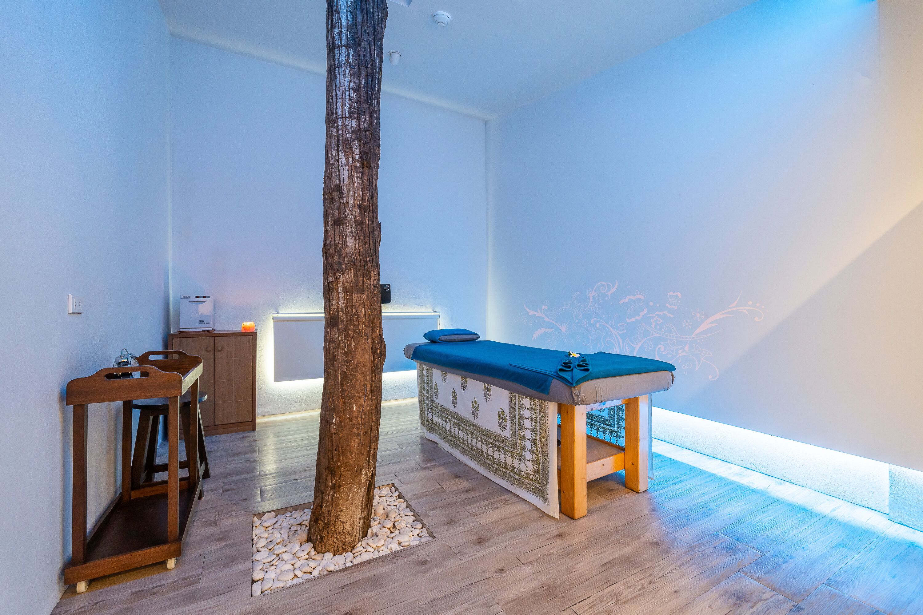 Una habitación con suelo de madera, un tronco de árbol grande, una mesa de masaje y un mueble pequeño.