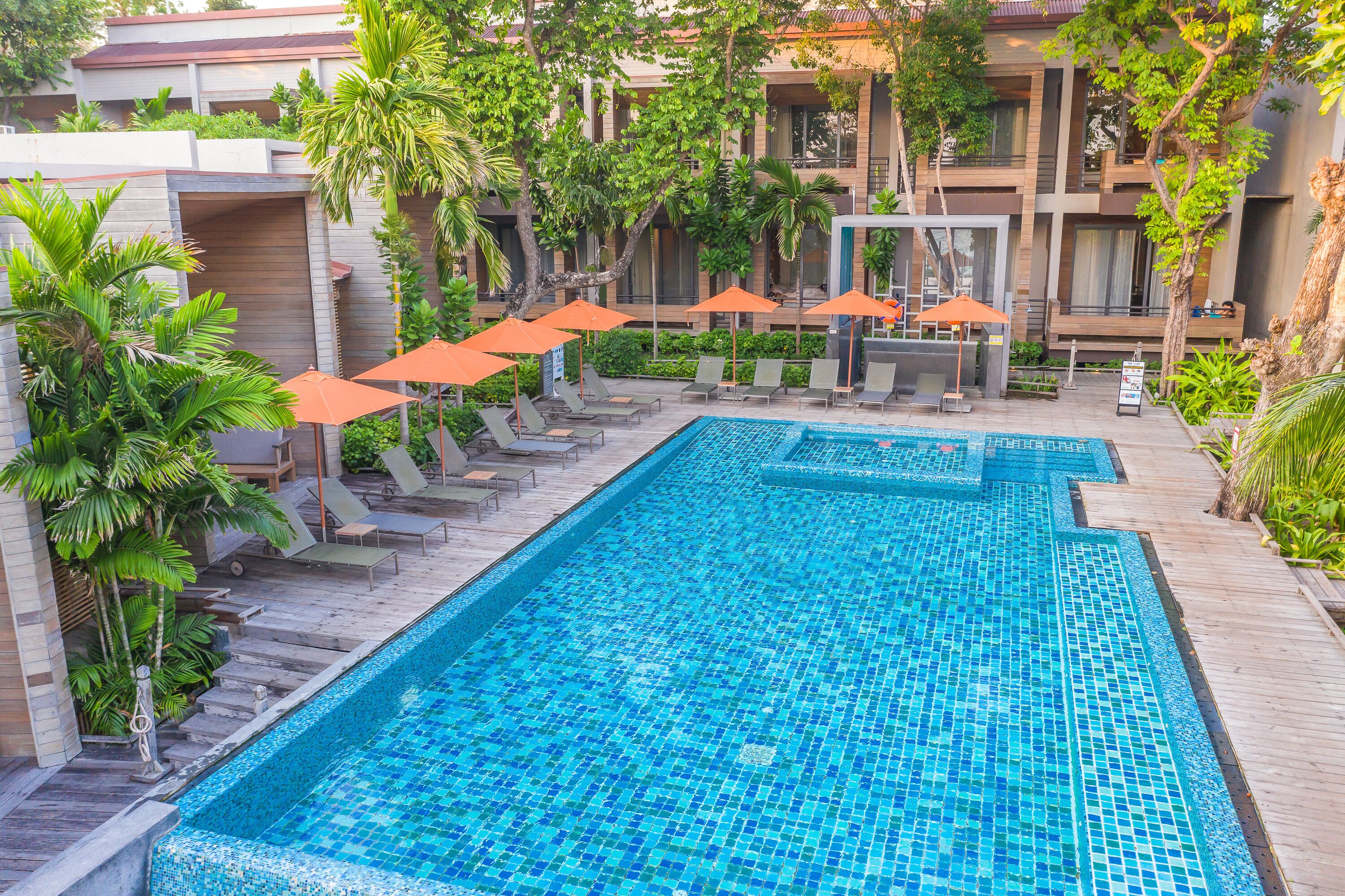 A rectangular pool with blue mosaic tiles, surrounded by lounge chairs and orange umbrellas, with a building in the background.