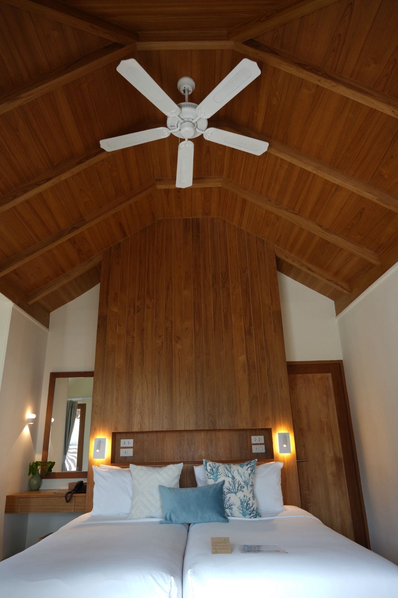 Une chambre avec un grand lit, des poutres apparentes en bois au plafond, un ventilateur de plafond, des lampes de chevet et une table de chevet ornée d’un vase et d’un téléphone.