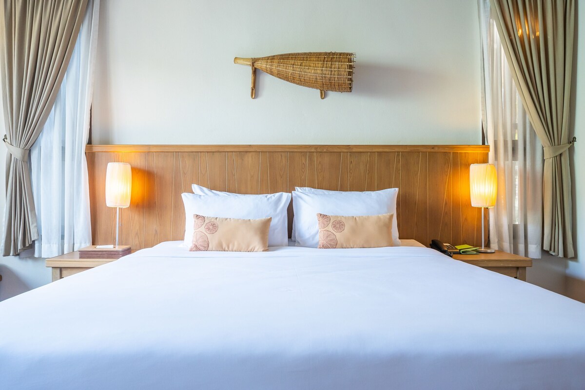 A hotel room with a wooden headboard, two bedside lamps, a bed with white and peach pillows, and a wall-mounted decorative item.
