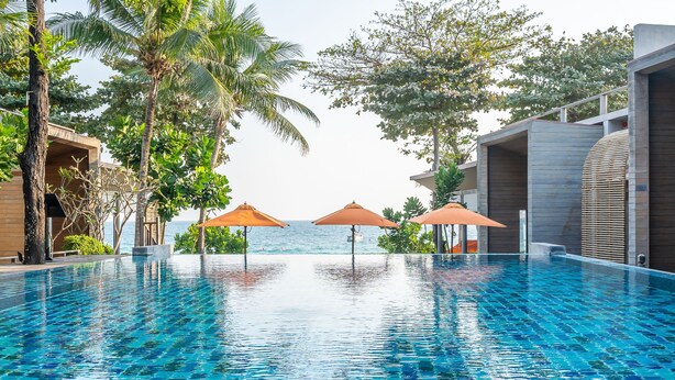 A swimming pool with clear blue water, surrounded by palm trees and umbrellas, with a view of the sea in the background.