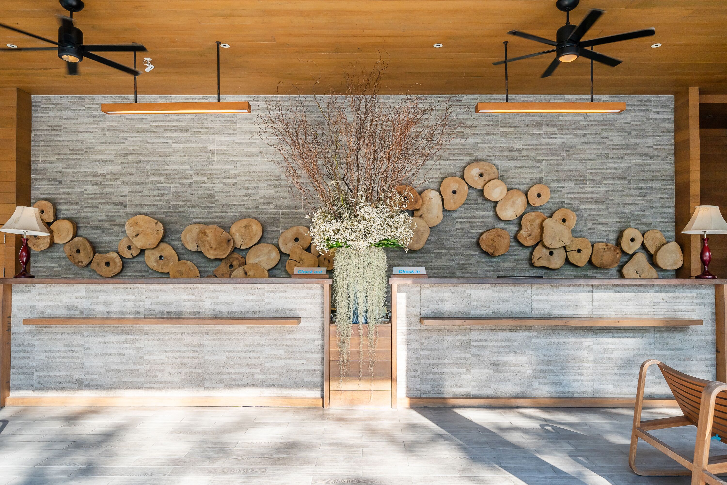 Un mostrador de recepción con una pared de ladrillo gris, troncos de madera y un jarrón con flores blancas.