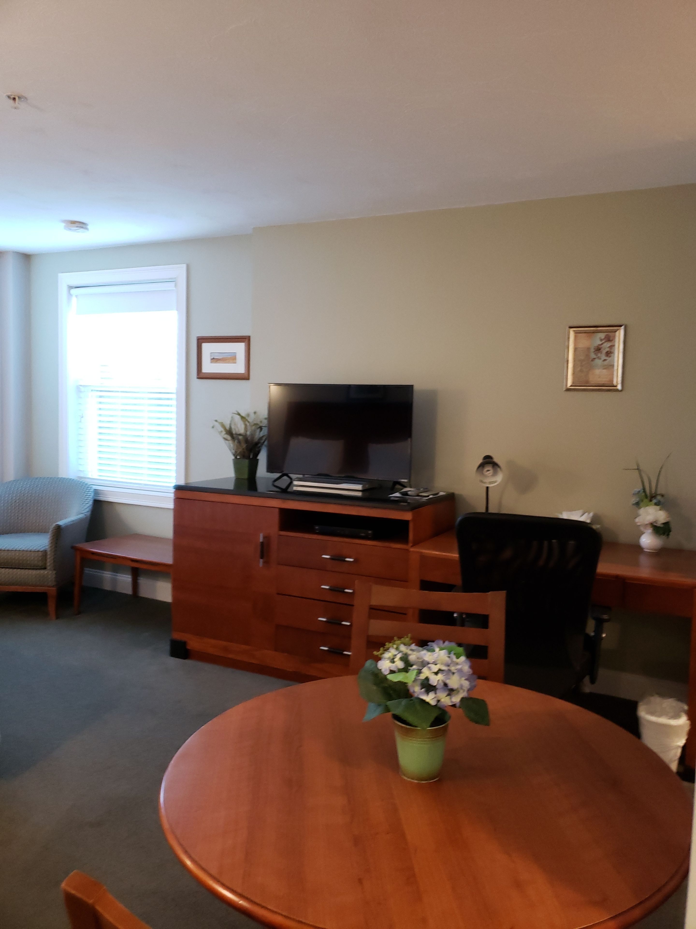 A living room with a wooden TV stand, a flat-screen TV, a dining table with a vase of flowers, a sofa, and a desk with a chair.