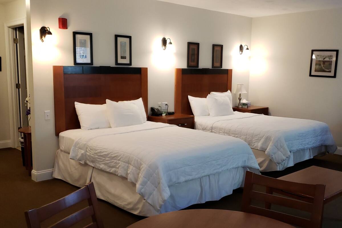 Quarto de hotel com duas camas, uma mesa de madeira redonda e uma cadeira.