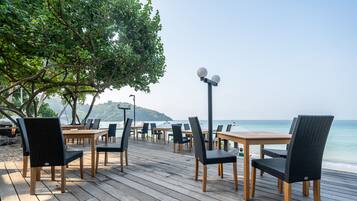 Un área de comedor al aire libre con mesas y sillas de madera, con vistas a una playa y al océano.