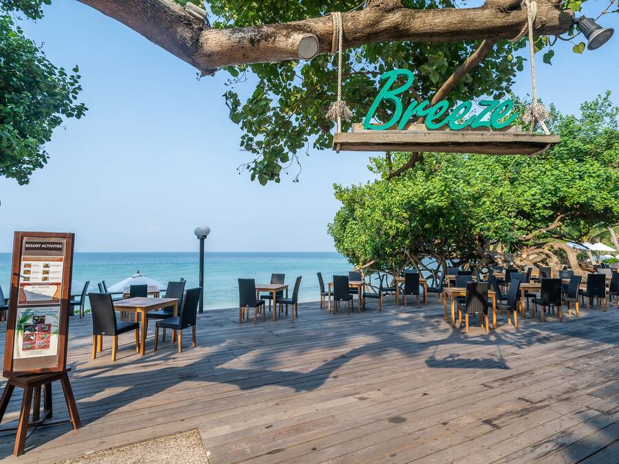 Una terraza de madera con mesas y sillas, un letrero colgado de un árbol y vista al mar.