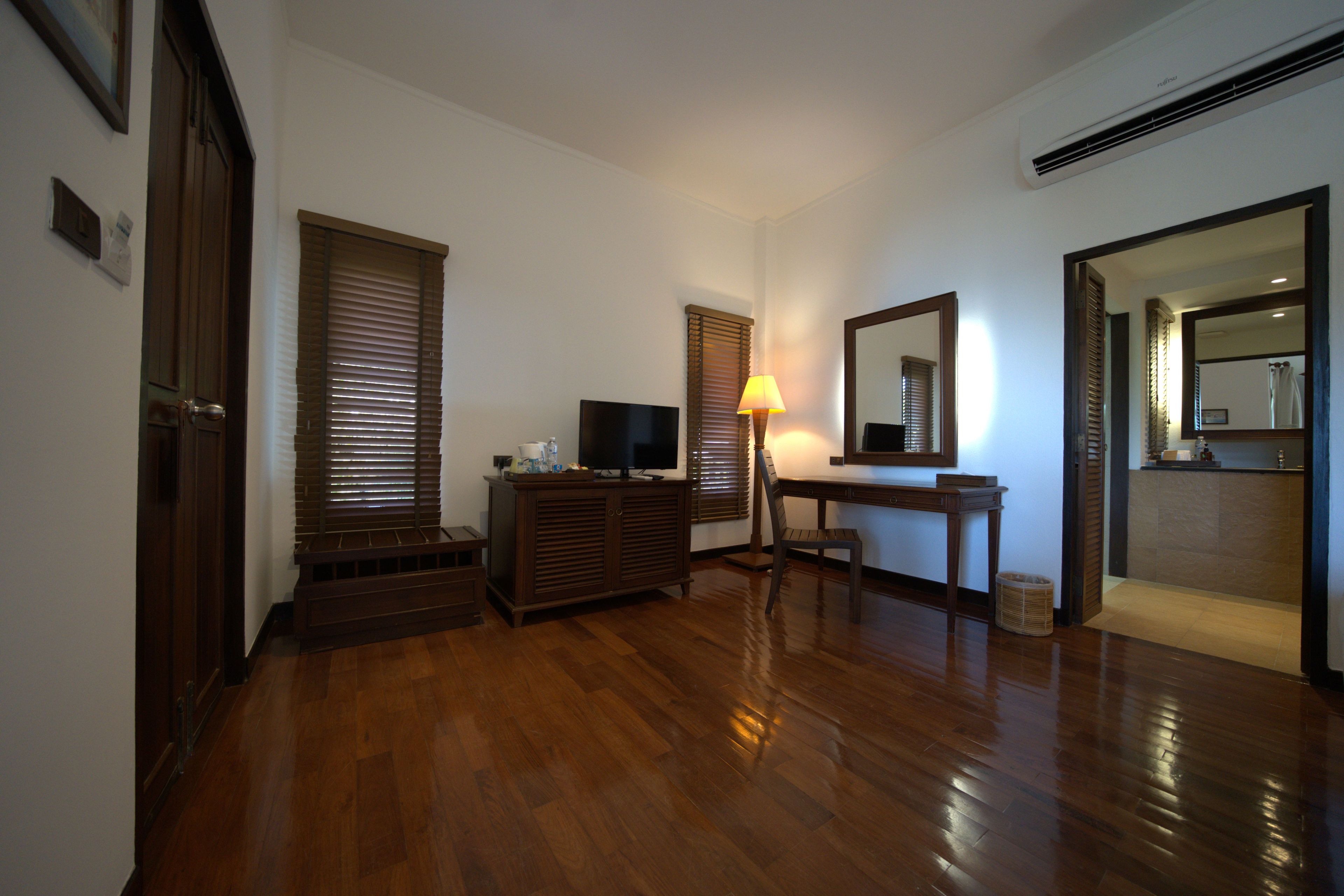 A room with wooden flooring, a television on a wooden cabinet, a desk with a lamp, and a mirror on the wall.